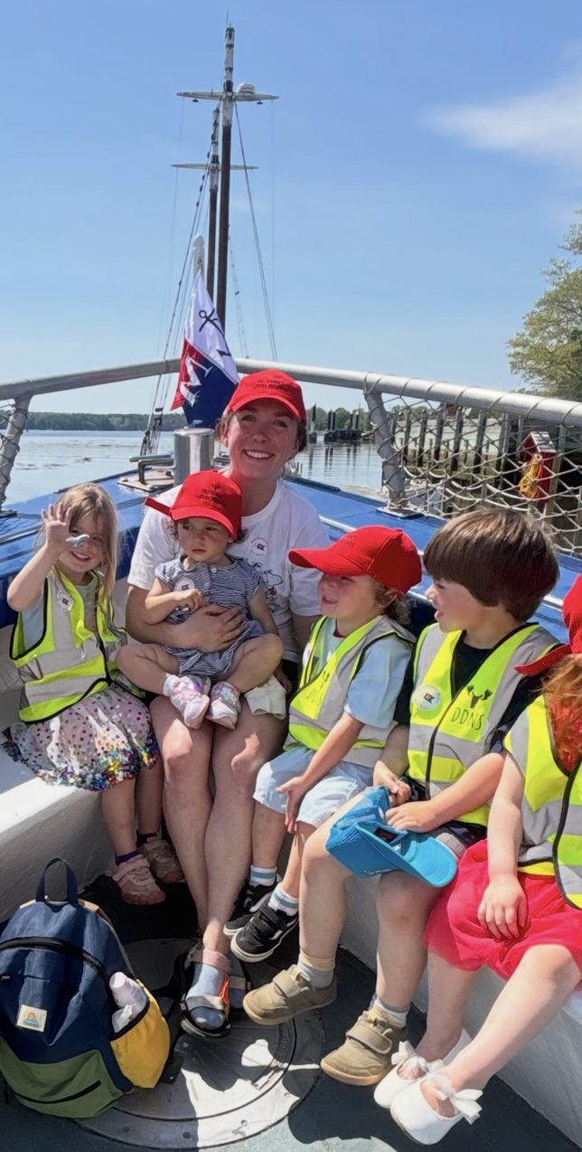 A teacher and children in matching red caps ride the bow of a schooner