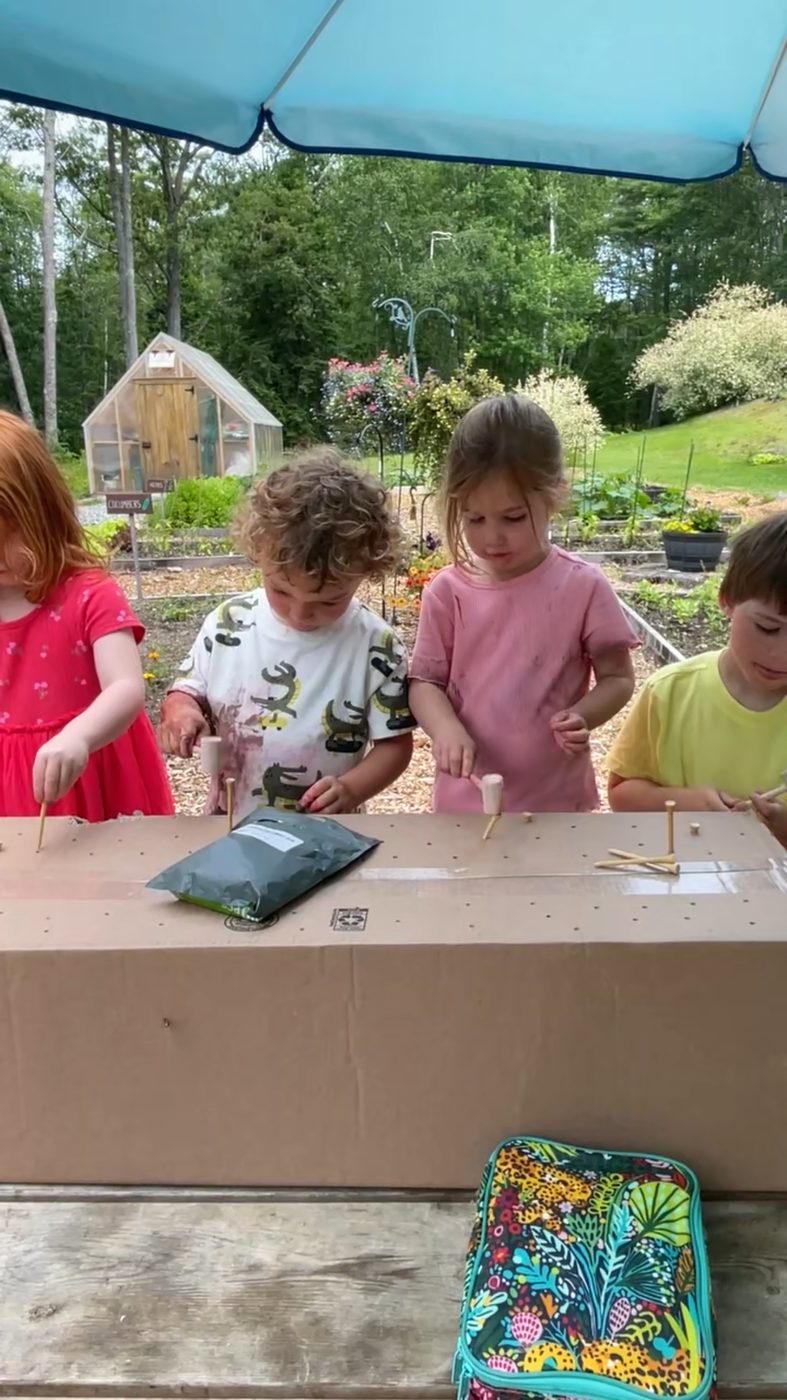 Children hammer wooden pegs at an outdoor workbench beside the gardens