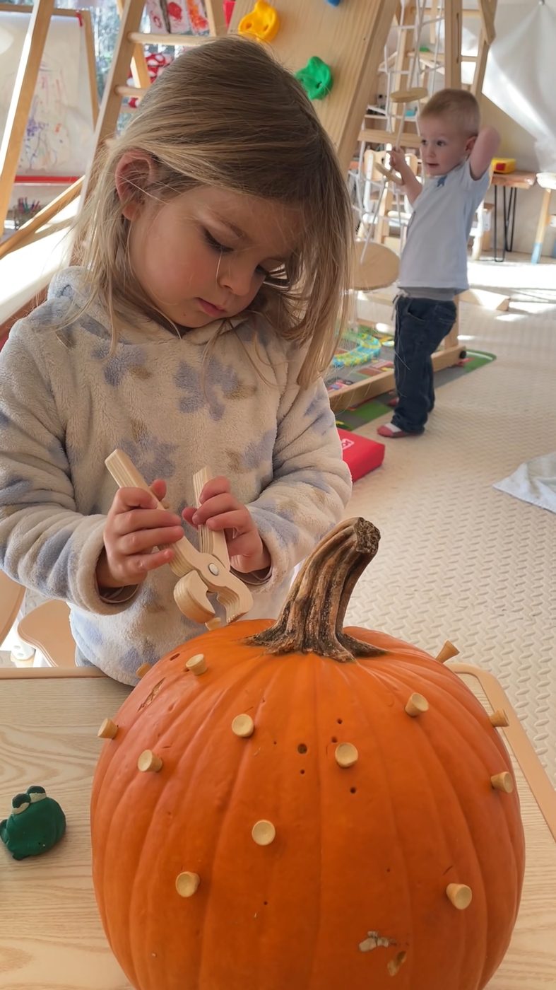 A child hammers wooden tees into a pumpkin with focused concentration