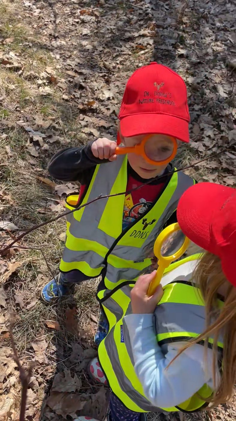 Two children in school hats and vests examine a branch with magnifying glasses