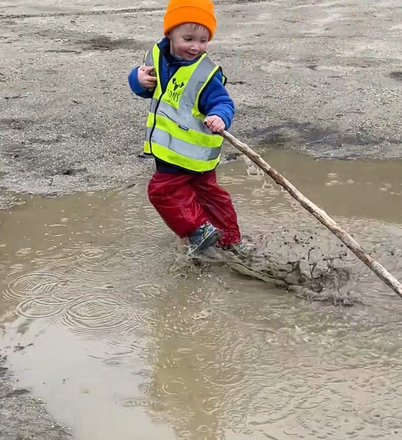 A child in an orange hat splashes through a mud puddle with a big stick