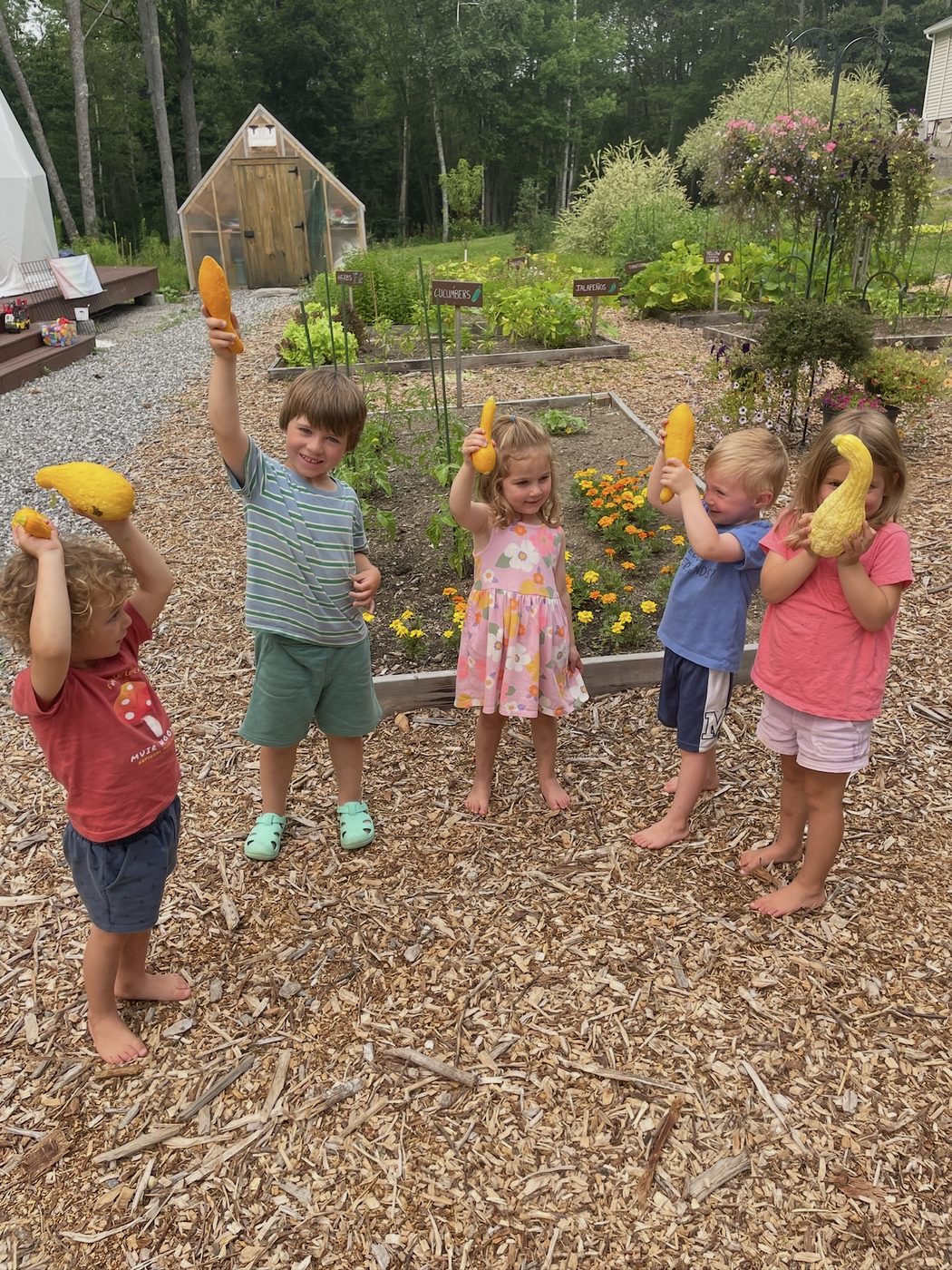 Children hold up yellow squash they grew, with the gardens, greenhouse, and geodesic dome behind them
